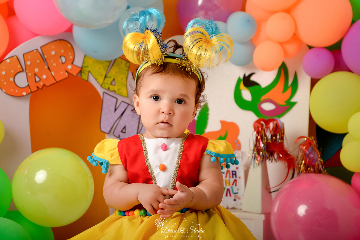 drica studio - ensaio fotográfico infantil - carnaval- xinguara - pará - bebê sentada em banquinho, usando vestido palhacinha e tiaras de coqueiro colorida-nome CAR NA VAL - máscara de carnaval -balões coloridos - fundo amarelo - painel branco