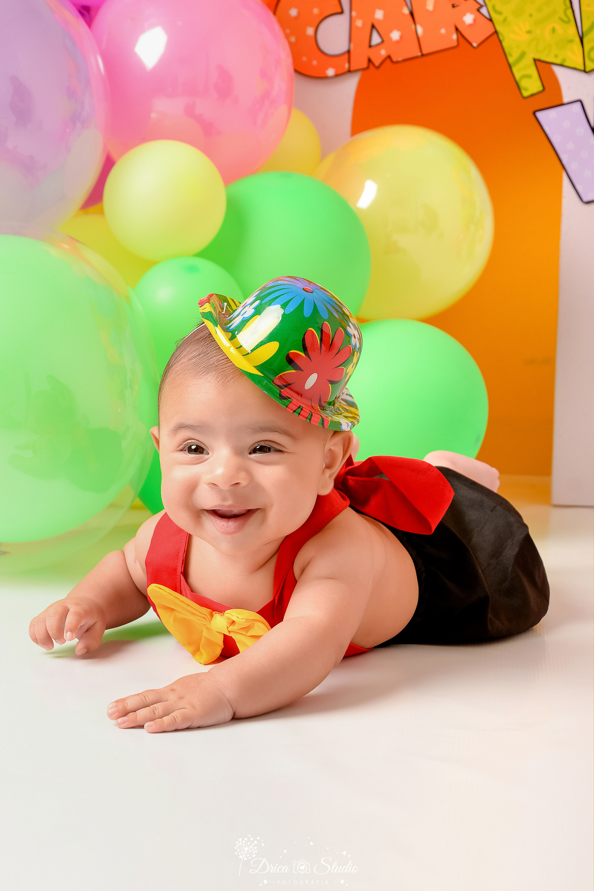 drica studio - ensaio fotográfico infantil - carnaval- xinguara - pará - bebê deitado de bruços, usando roupinha de palhaço e chapéu colorido - balões verde e amarelo - fundo amarelo- painel lilás