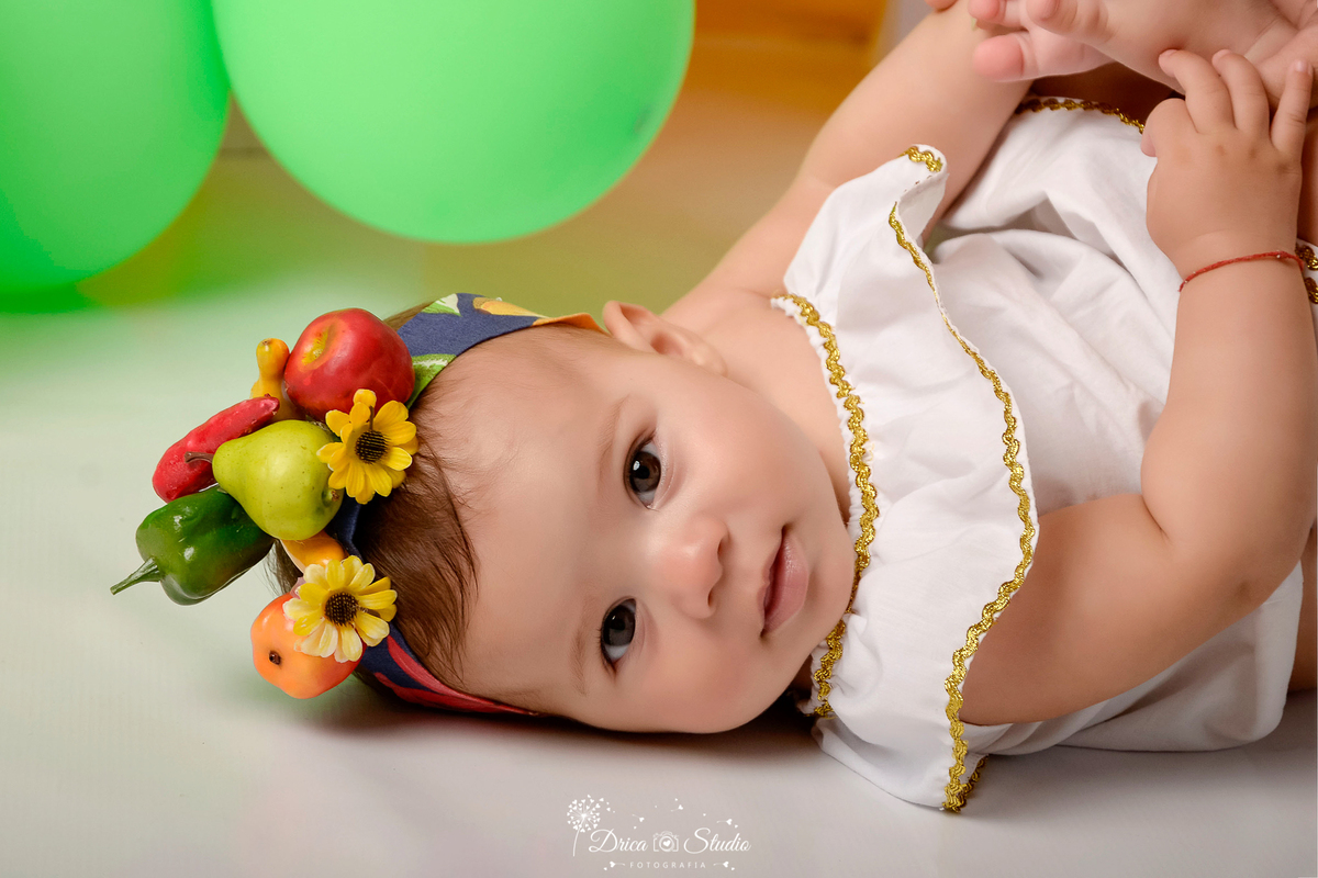 drica studio - ensaio fotográfico infantil - carnaval- xinguara - pará - bebê deitada no chão usando roupa de carmem miranda e frutinhas na cabeça - balões verdes - fundo amarelo - piso branco