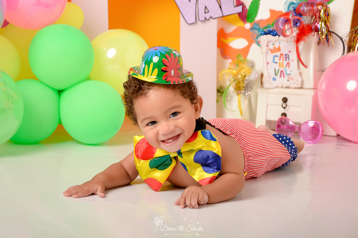 drica studio - ensaio fotográfico infantil - carnaval- xinguara - pará - bebê deitado de bruços, usando calça de palhoço com suspensório e gravata borboleta - chapéu colorido - balões coloridos - máscara de carnaval 