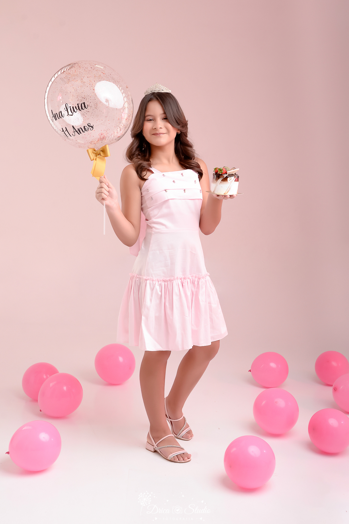 Drica studio - ensaio fotografico infantil- xinguara - pará - Retrato infantil feminino de 11 anos com tiara e vestido rosa, segurando balão personalizado “Ana Lívia 11 anos” e bolo de aniversário em estúdio com fundo rosa.