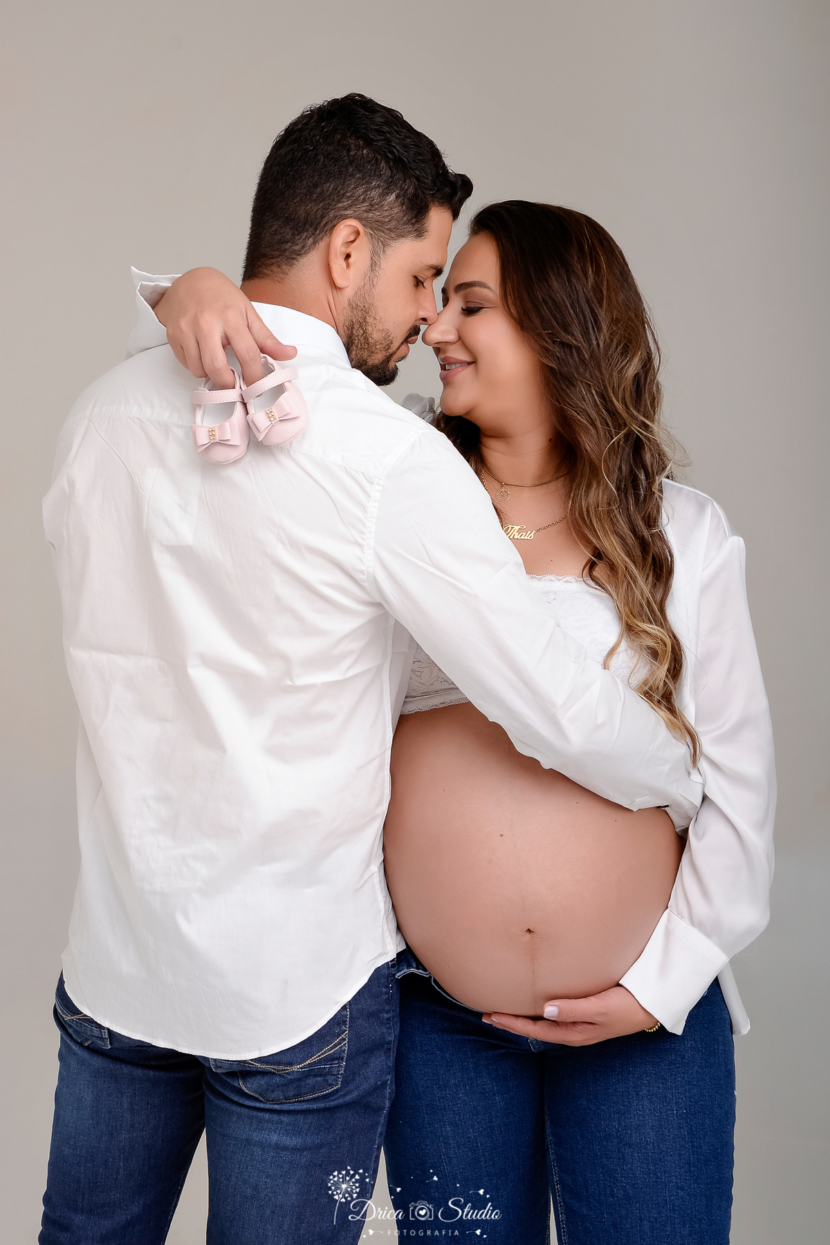 drica studio - ensaio fotografico gestação - xinguara - pará - casal posam para fotos gestacional - homem de costa, em pé ao lada da esposa abraçando-a e trocando olhares- ambos vestem branco - fundo neutro