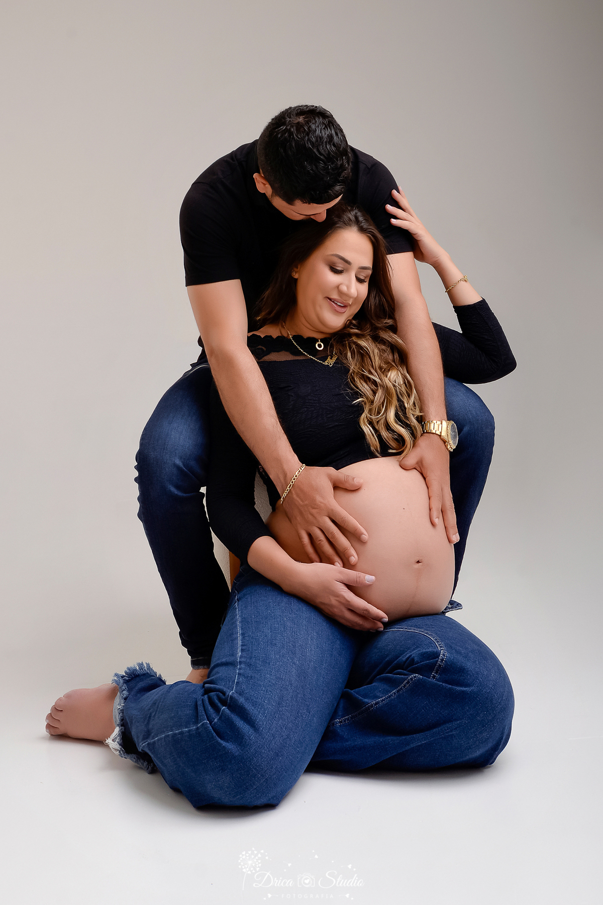 drica studio - ensaio fotografico gestação - xinguara - pará - casal pasam para foto gestacional - esposa sentado em banqueta e esposa sentada no chão - esposo com as mãos sobre a barriga da gestante - ambos veste camisa e blusa preta e calça jean 