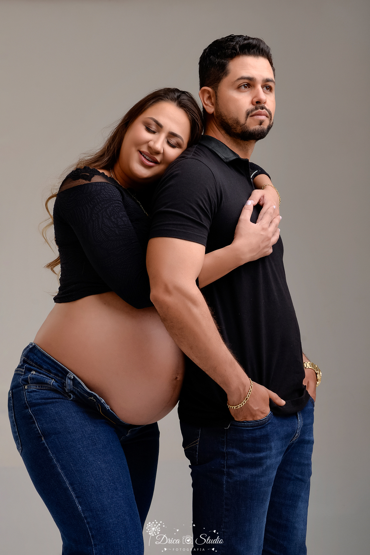 drica studio - ensaio fotografico gestação - xinguara - pará -casal posam para foto gestacional, mulher abraça o esposo por tras, encostando o rosto no ombro do esposo-mulher veste crope preto e deixa a barriga amostra - ambos veste caça jean-fundo neutro