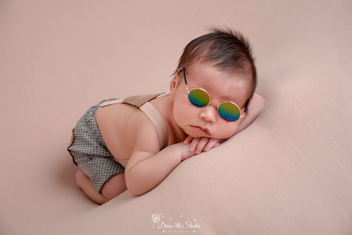 drica studio-ensaio fotografico recém-nascido- newborn-xinguara - pará - bebê recém-nascido apoiando o rostinho nas mãos - usando short com suspensório sem camisa e óculos redondo de sol - fundo neutro