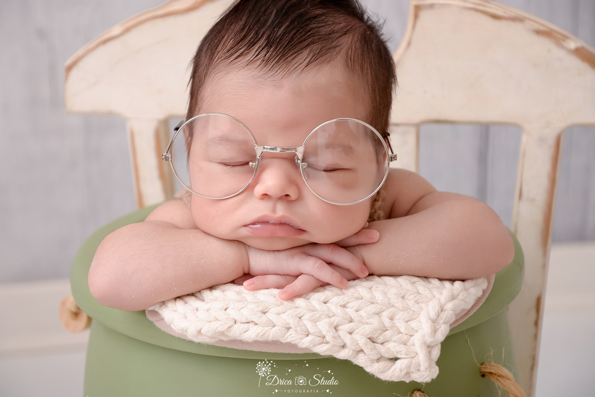 rica studio-ensaio fotografico recém-nascido- newborn-xinguara - pará -bebê recém-nascido usando óculos oversized dormindo dentro de balde de madeira verde, acochoado com manta bege clara de trico- o balde está sobre cadeira de madeira