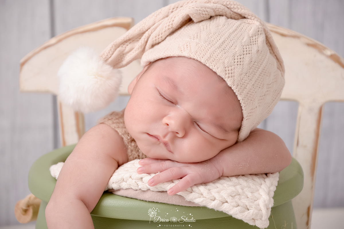 drica studio-ensaio fotografico recém-nascido- newborn-xinguara - pará - bebê recém-nascido dormindo dentro de baldinho de madeira verde, acochoado com manta bege, ele sua toca bege com pompo branco na ponta e apoia o rosto sobre a mão - fundo claro