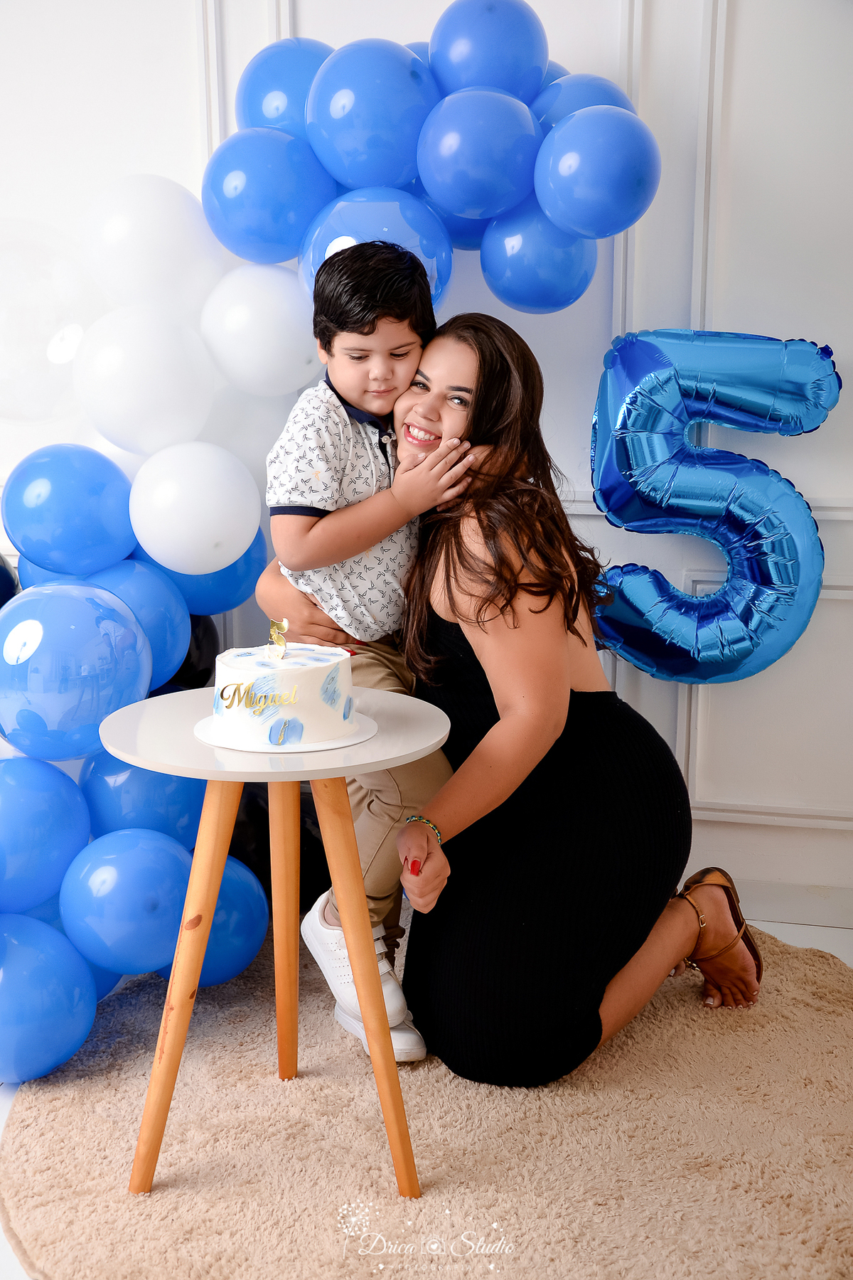 drica studio - ensaio fotografico infantil - 5 anos - xinguara - pará - mãe abraçada ao filho, ele a toca o rosto com a mão carinhamente - balão azul - balão número 5 - fundo neutro - bolo branco com mancha azuis sobre mesinha branca