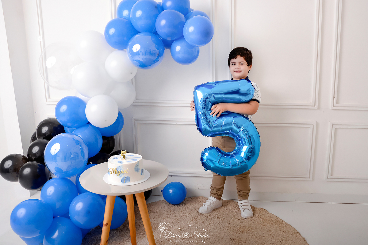 drica studio - ensaio fotografico infantil - 5 anos - xinguara - pará - menino segurando numero 5 - balões azul, preto e branco - bolo branco sobre uma mesinha clara com perna de madeira - fundo neutro - tapete redondo
