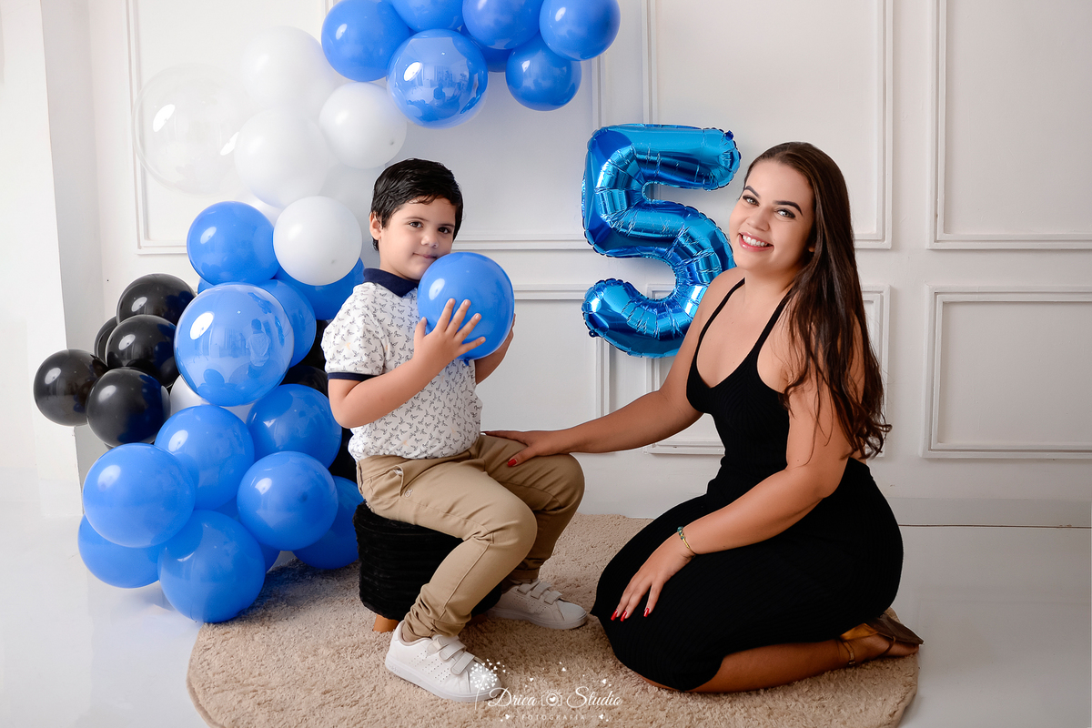 drica studio - ensaio fotografico infantil - 5 anos - xinguara - pará - menino sentado em pulf de frente com a mãe - ele veste camisa branca e calça bege, ela vestido preto - balões azul, preto e branco - balão número 5 - fundo branco com boiserie