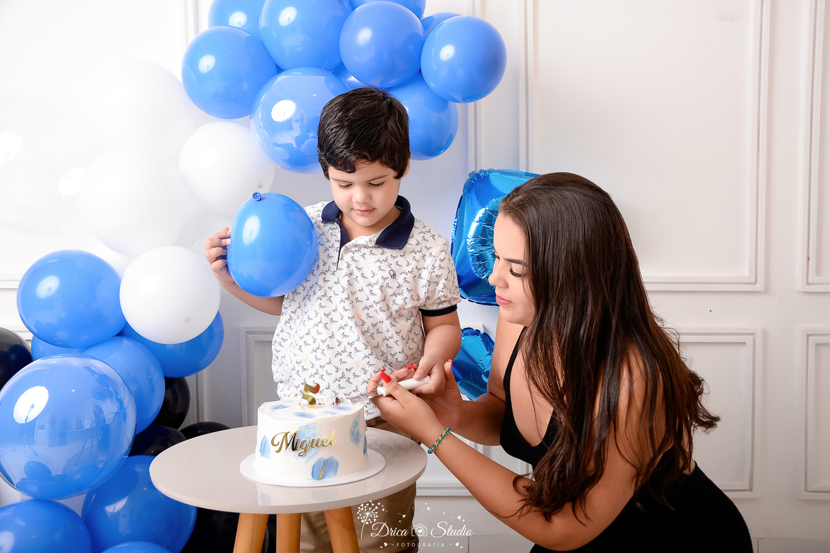 drica studio - ensaio fotografico infantil - 5 anos - xinguara - pará - menino cortando o bolo com auxilio da mãe - ele veste camisa branco com detalhes azuis e calça bege, ela veste vestido preto - balões azuis, preto e branco - fundo branco