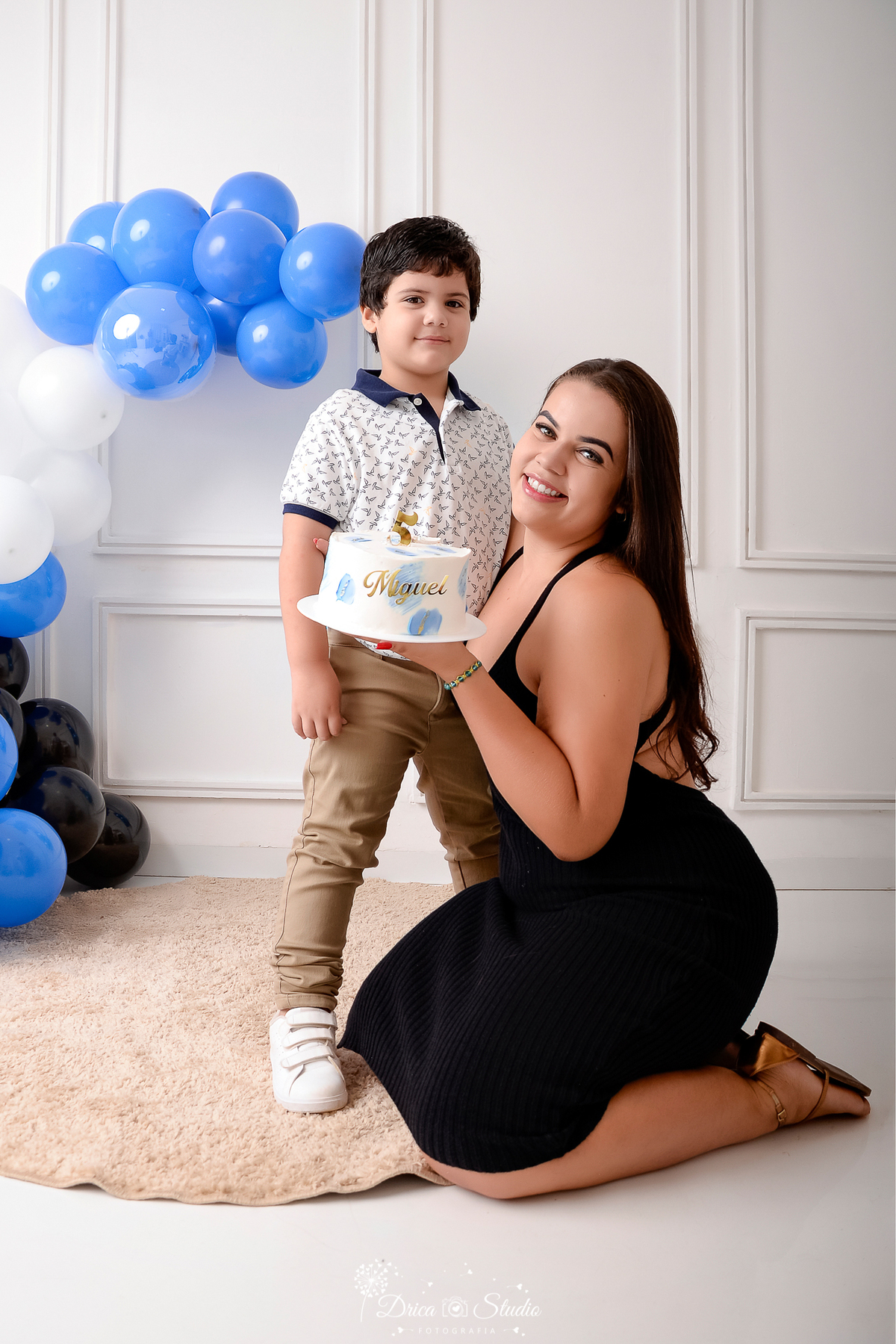 drica studio - ensaio fotografico infantil - 5 anos - xinguara - pará - mãe segurando bolo de aniversário do filho, ela veste vestido de alçinha preto-ele camisa branca e calça bege - fundo branco com boiserie - balão azul,preto e branco