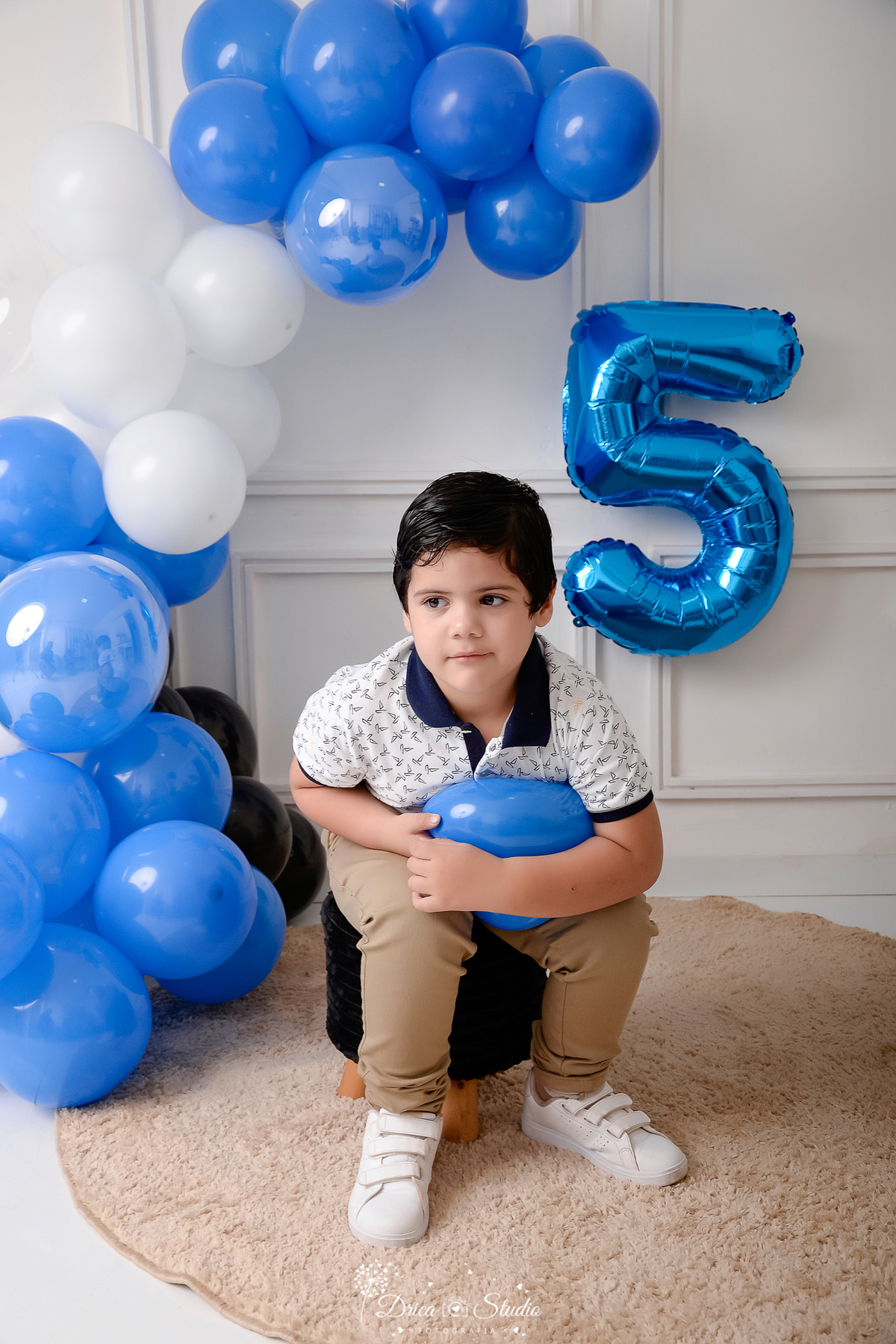 drica studio - ensaio fotografico infantil - 5 anos - xinguara - pará - balões azuis, branco e preto - balão número 5 - menino sentado em pul preto, segurando balão - fundo branco de boiserie