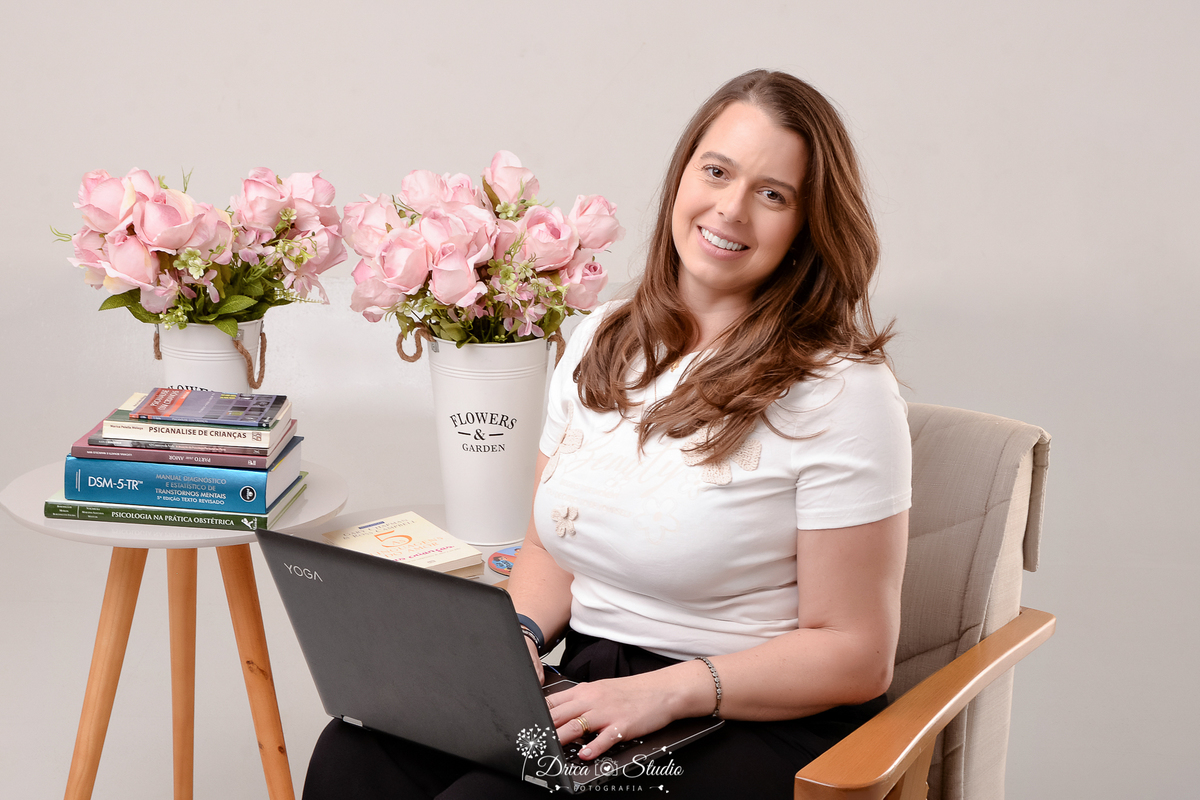 drica studio - ensaio fotografico profissional - psicologia infantil - xinguara - pará-Psicóloga em retrato corporativo sentada com notebook, livros técnicos e DSM-5-TR ao lado- ela veste blusa branca e calça preta - fundo branco