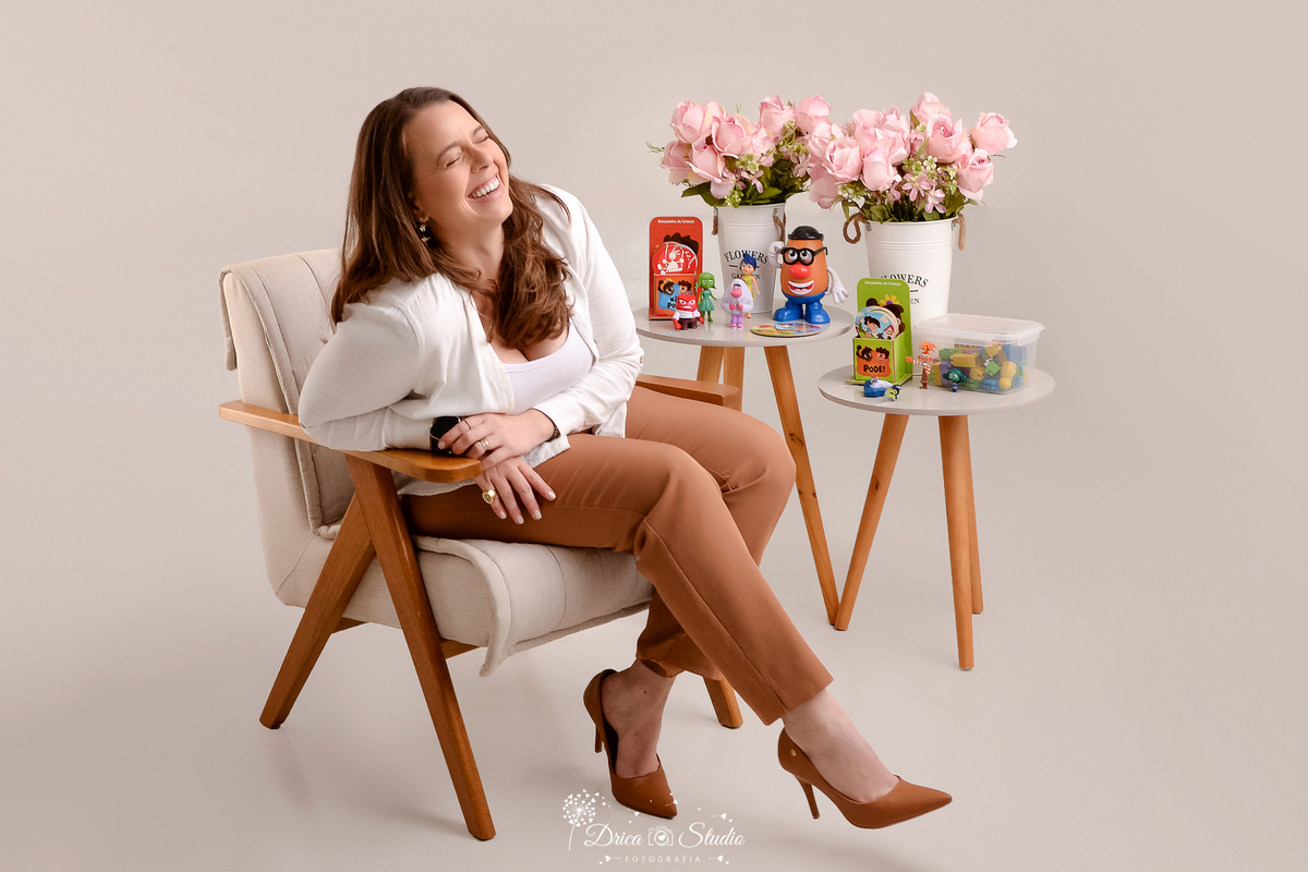 Psicóloga infantil em ensaio drica studio - ensaio fotografico profissional - psicologia infantil - xinguara - pará-profissional sentada em poltrona, sorrindo, com brinquedos terapêuticos e flores ao fundo.