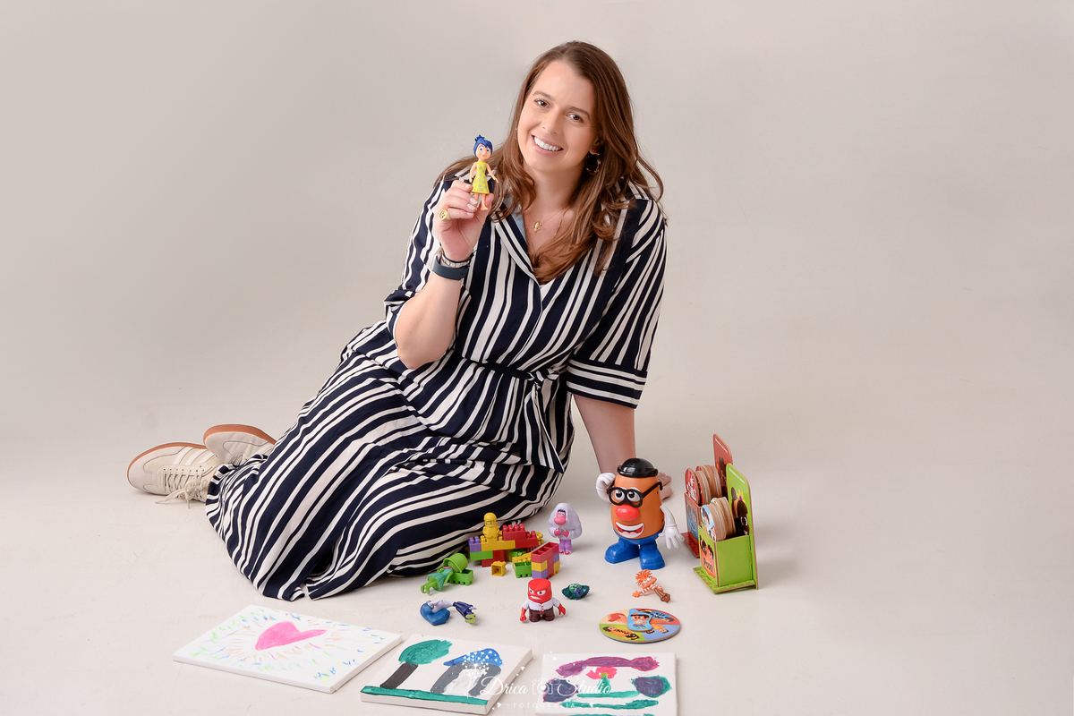 drica studio - ensaio fotografico profissional - psicologia infantil - xinguara - pará- Psicóloga sentada no chão com brinquedos e desenhos infantis, segurando personagem lúdico- veste vestido longo branco com listras pretas - fundo branco