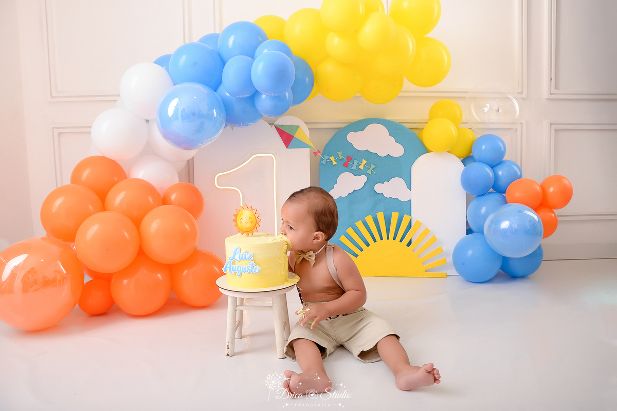 drica studio-ensaio fotografico infantil - xinguara - pará - smash the cake - minha primeira volta ao sol - hum de led - solzinho - bebê sentado no chã mordendo bolo - balões laranja, branco, amarelo e azul - pipa colorida - fundo branco com boiserie