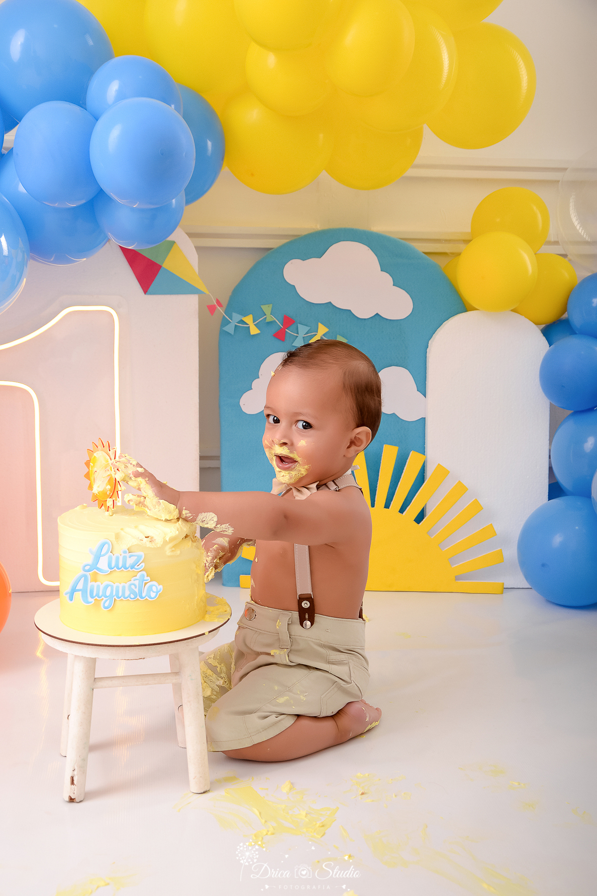 drica studio-ensaio fotografico infantil - xinguara - pará - smash the cake - minha primeira volta ao sol - hum de led - solzinho - bebê sentado no chão amassando bolo comas mãos, ele veste short bege com suspensório e gravatinha borboleta - painel azul