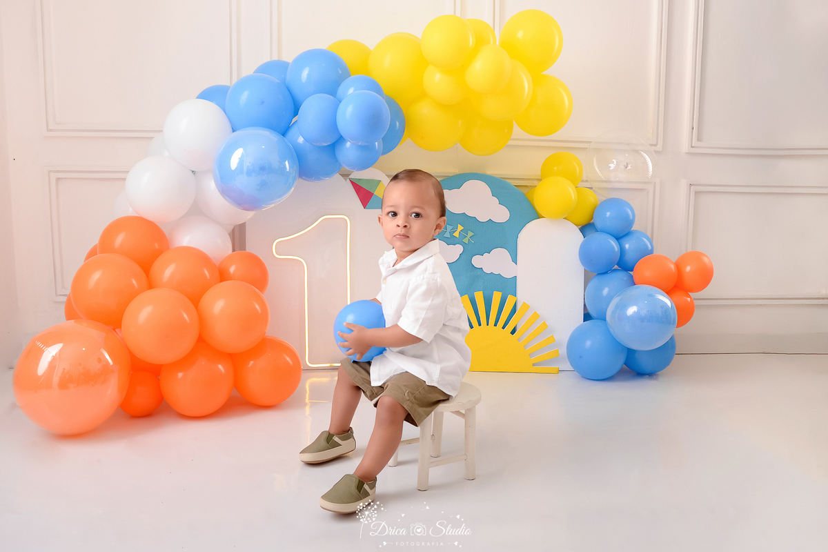 drica studio-ensaio fotografico infantil - xinguara - pará - smash the cake - minha primeira volta ao sol - hum de led - solzinho - balões coloridos - bebê sentado em banquinho branco , ele veste camisa branca, short marrom e tênis marrom - fundo branco