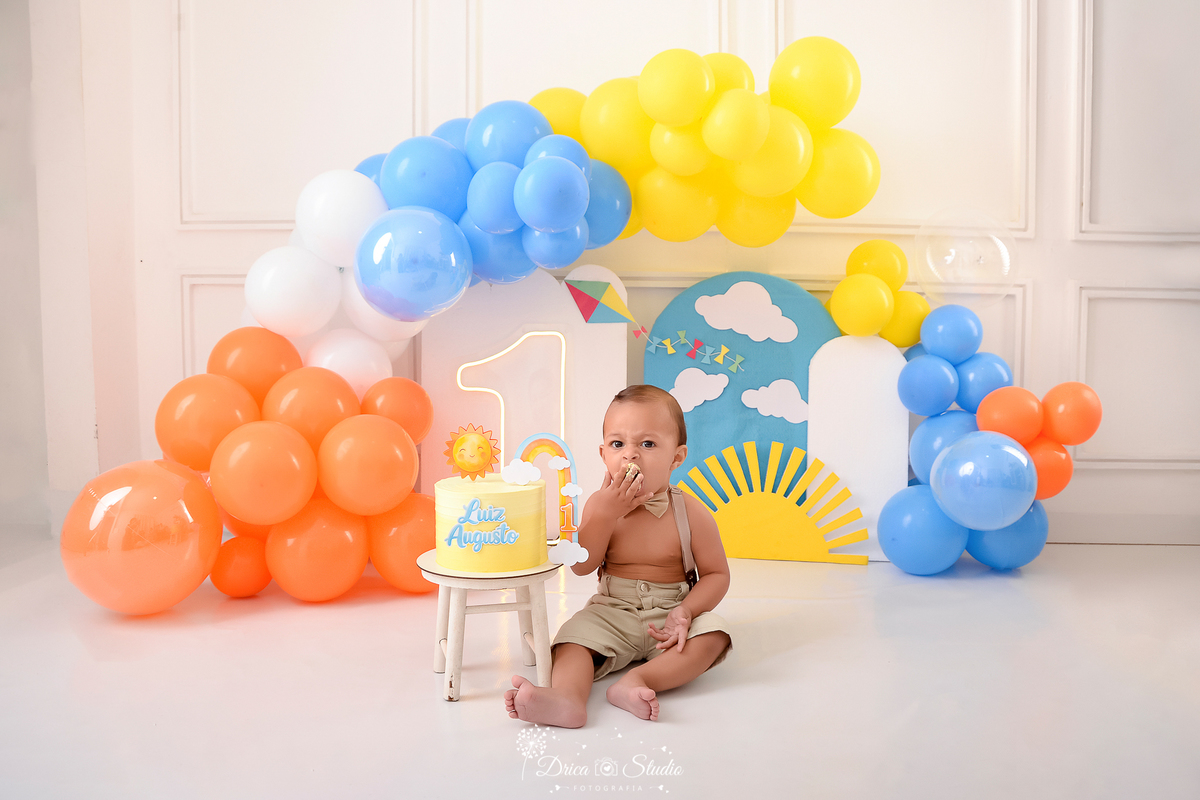drica studio-ensaio fotografico infantil - xinguara - pará - smash the cake - minha primeira volta ao sol - hum de led - solzinho - bebê sentado no chão ao lado do bolo que está sobre um banquinho branco - balões amarelo, laranja, azul e branco 