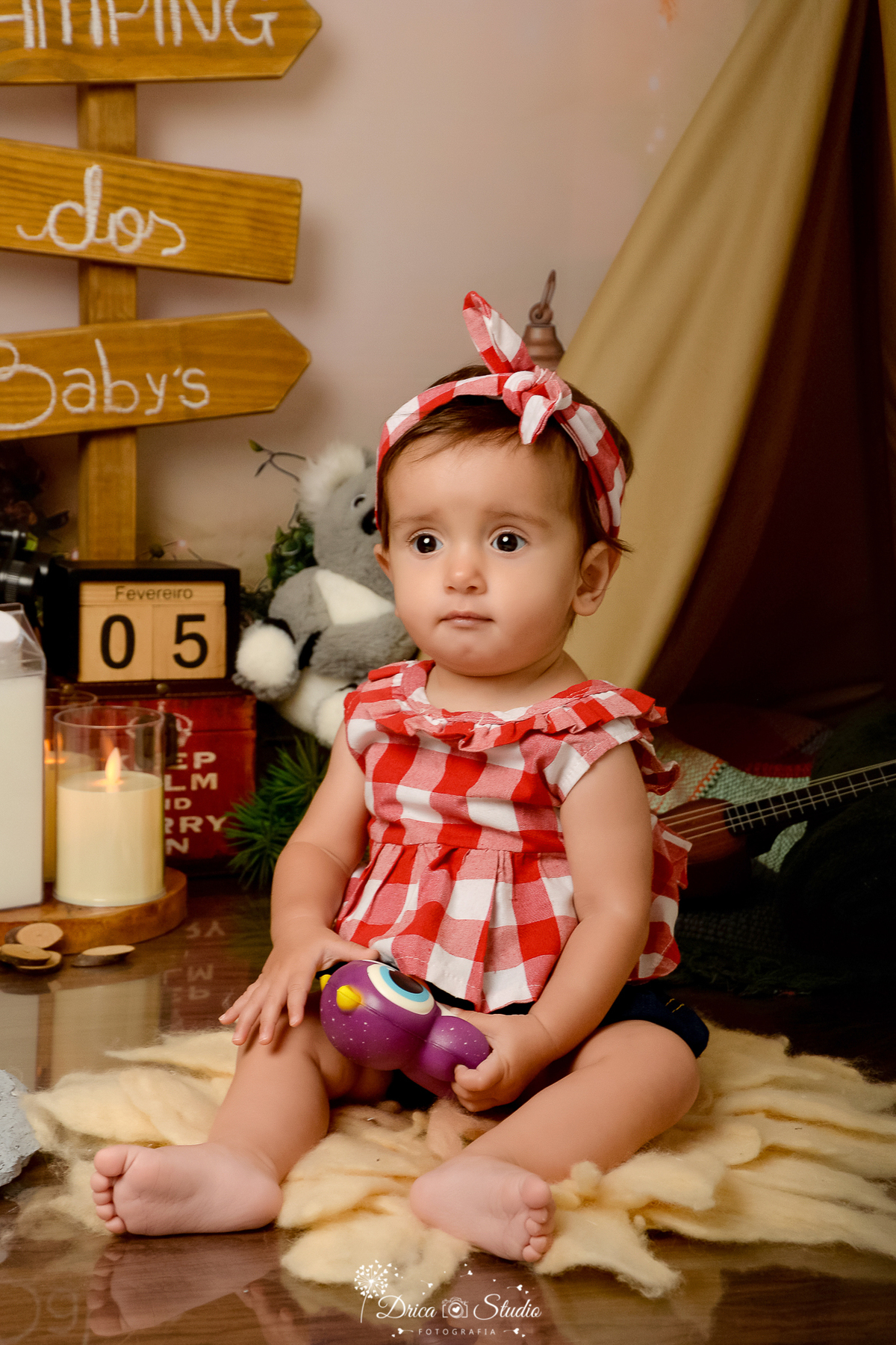 drica studio-ensaio fotografico infantil - acampamento infantil - xinguara - pará - bebê sentada no chão sobre tapete felpudo, ela veste blusa e faxinha xadrez - velas fake - cabaninha - violãozinho