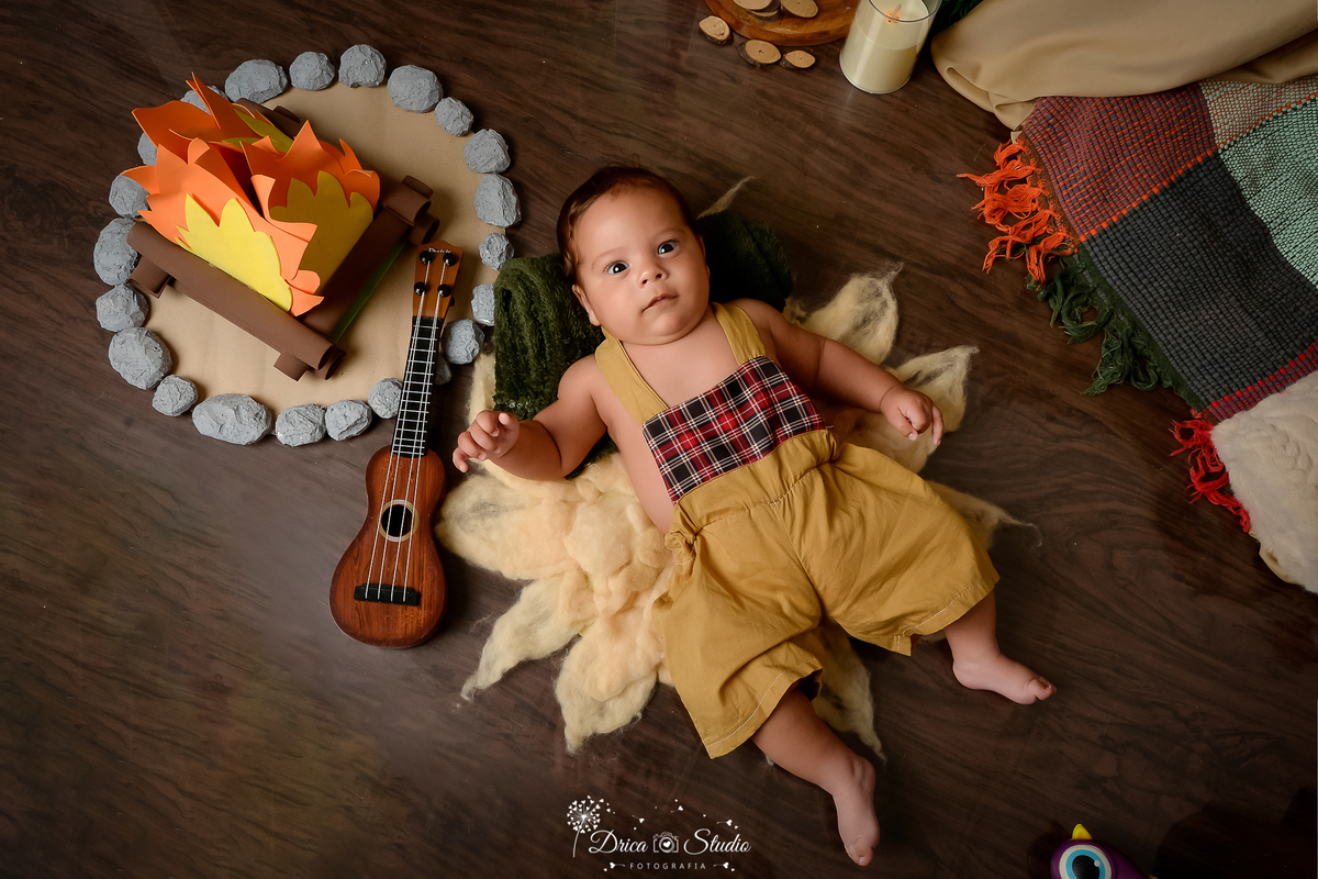 drica studio-ensaio fotografico infantil - acampamento infantil - xinguara - pará -bebê deitado sobre tapete felpudo, ele usa macaquinho mostada com estampa xadrez - fogueira de eva - violão - piso amadeirado - cabaninha 