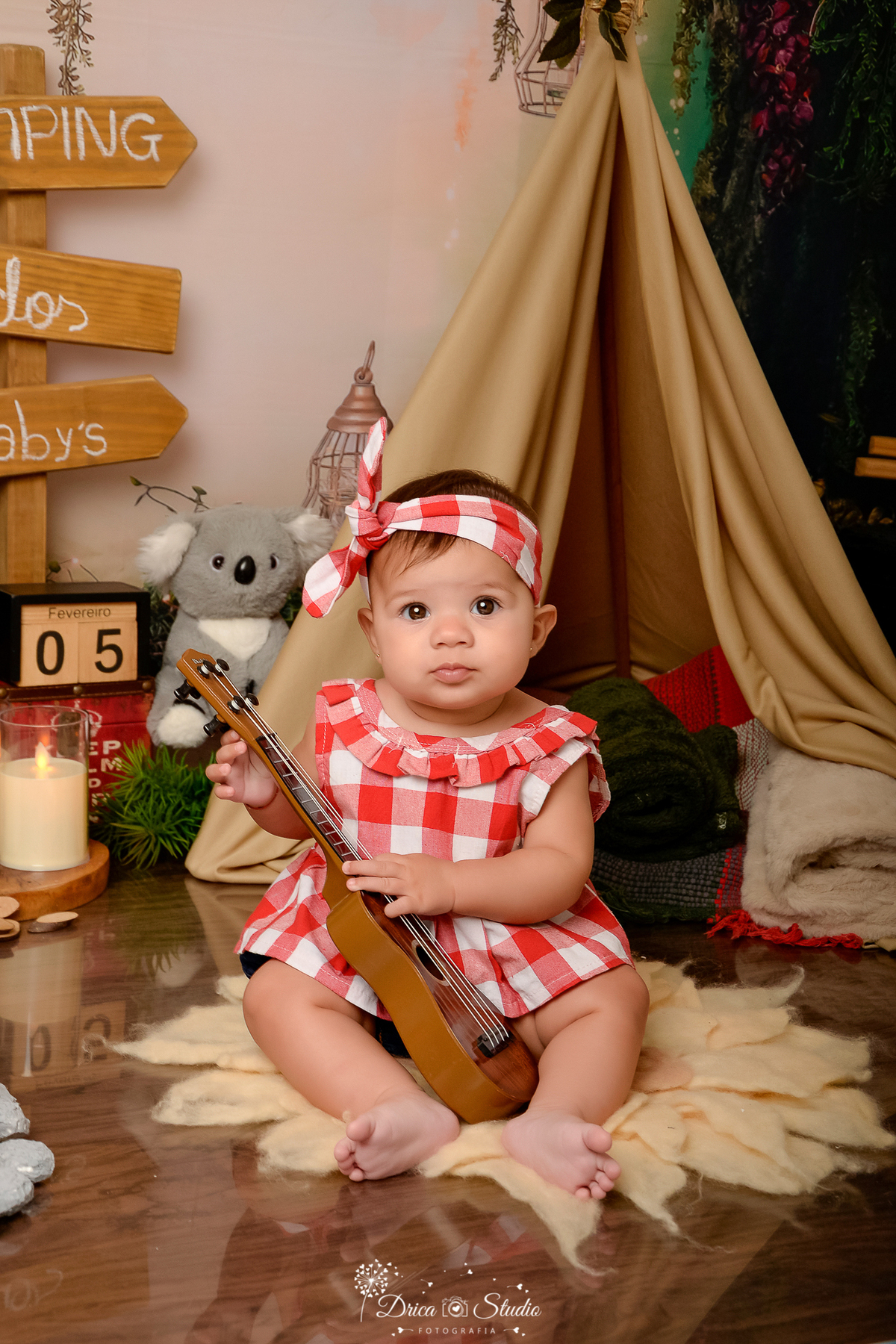 drica studio-ensaio fotografico infantil - acampamento infantil - xinguara - pará - bebê sentado segurando violão - cabaninha bege - vela de led - piso amadeirado - bichinho de pelúcia - plaquinha de indicação - manta verde escuro