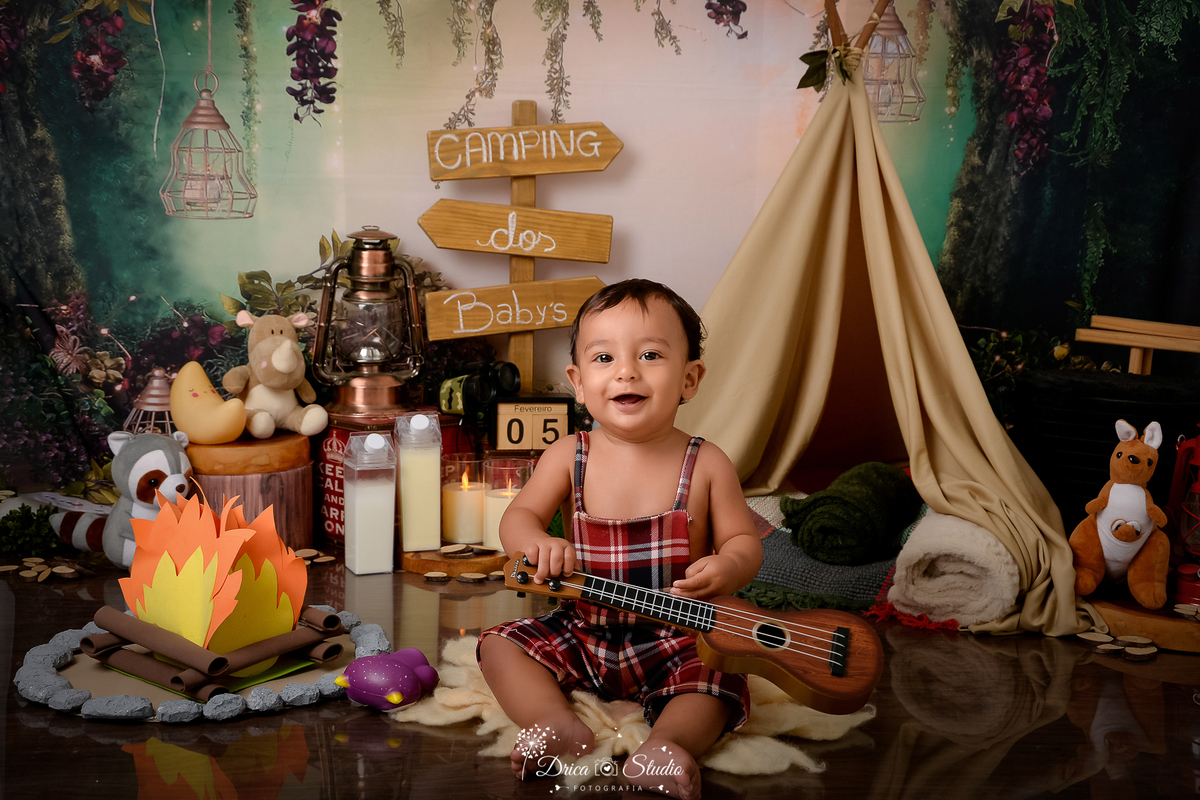 drica studio-ensaio fotografico infantil - acampamento infantil - xinguara - pará -bebê sentado no chão segurando violão vestido com macaquinho xadrez sem camisa- cabaninha - bichinhos de pelúcia - lampião - fogueirinha de eva - 