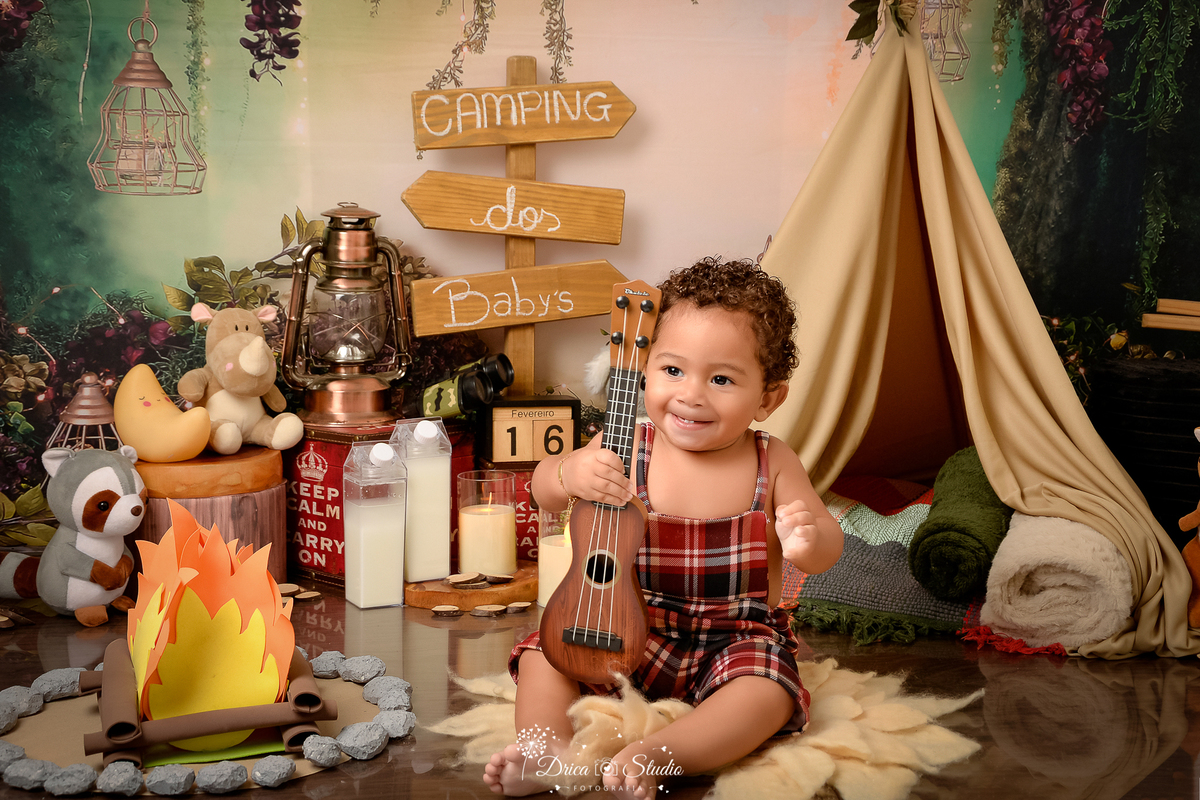 drica studio-ensaio fotografico infantil - acampamento infantil - xinguara - pará -bebê sentado no chão segurando violão - plaquinha com setas de indicação - fundo tema floresta - bichinhos de pelucia-lampião-cabaninha 