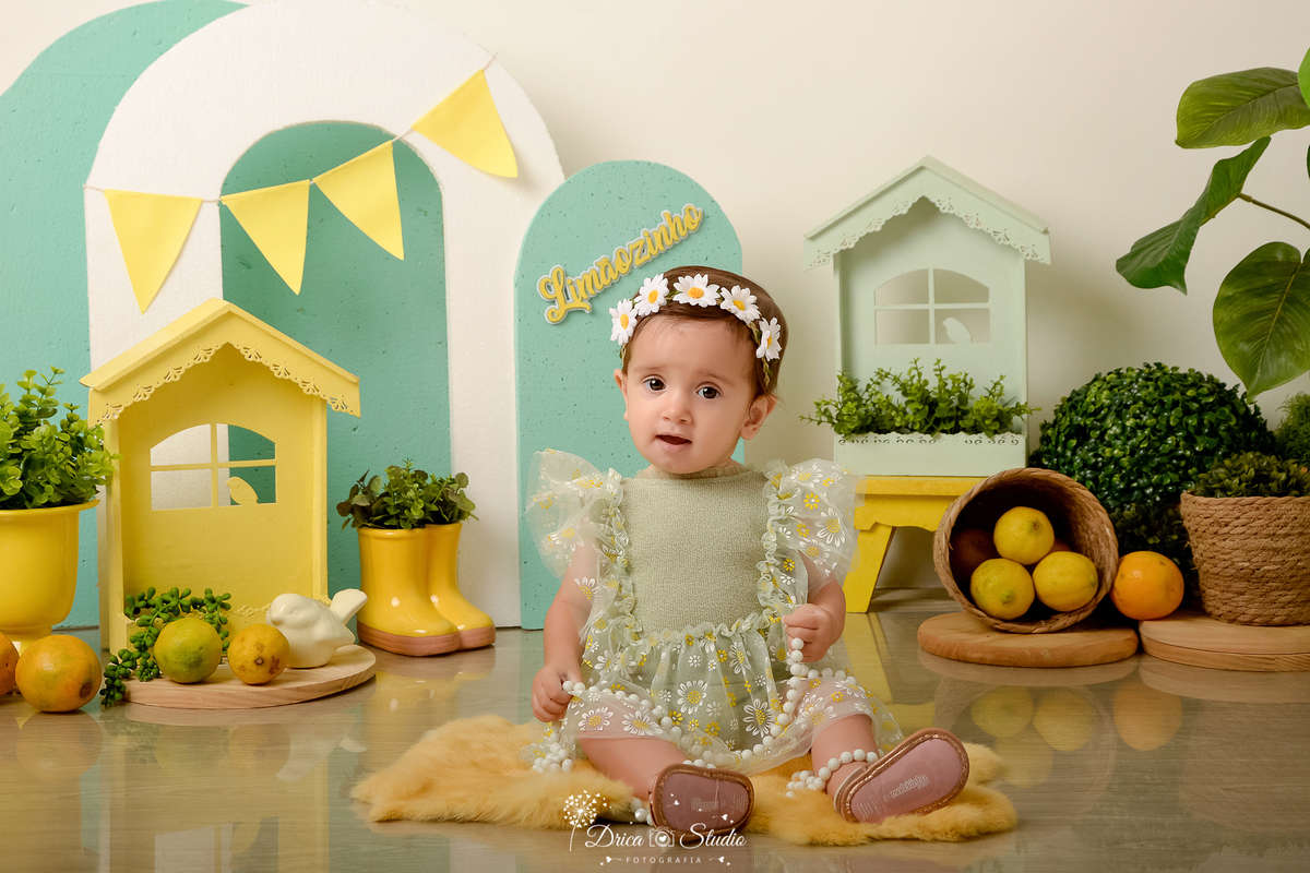 drica studio-ensaio fotográfico infantil - acompanhamento mensal - tema limãozinho -bebê sentada sobre tapete felpudo amarelo, veste macaquinho verde claro, casinhas verde e amarela - enfeite botinha amarela - limões - cestos de palha com plantas 