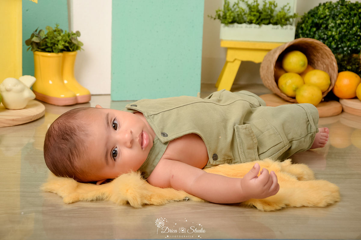 drica studio-ensaio fotográfico infantil - acompanhamento mensal - tema limãozinho - bebê deitado sobre tapete felpudo amarelo - macaquinho verde oliva - limões dentro de cestinhos de palha - botinha amarela com planta - banquinho amarelo - painel verde -