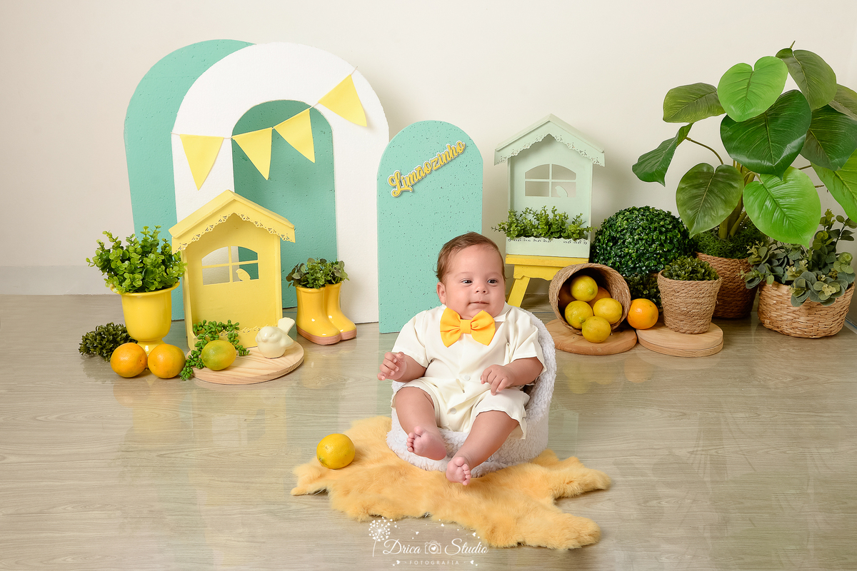 drica studio-ensaio fotográfico infantil - acompanhamento mensal - tema limãozinho - bebê vestindo macaquinho branco sentado em boltrona branca - gravatinha borboleta amarela - painel verde com bandeirolas amarela - casinhas verde e amarela - fundo neutro