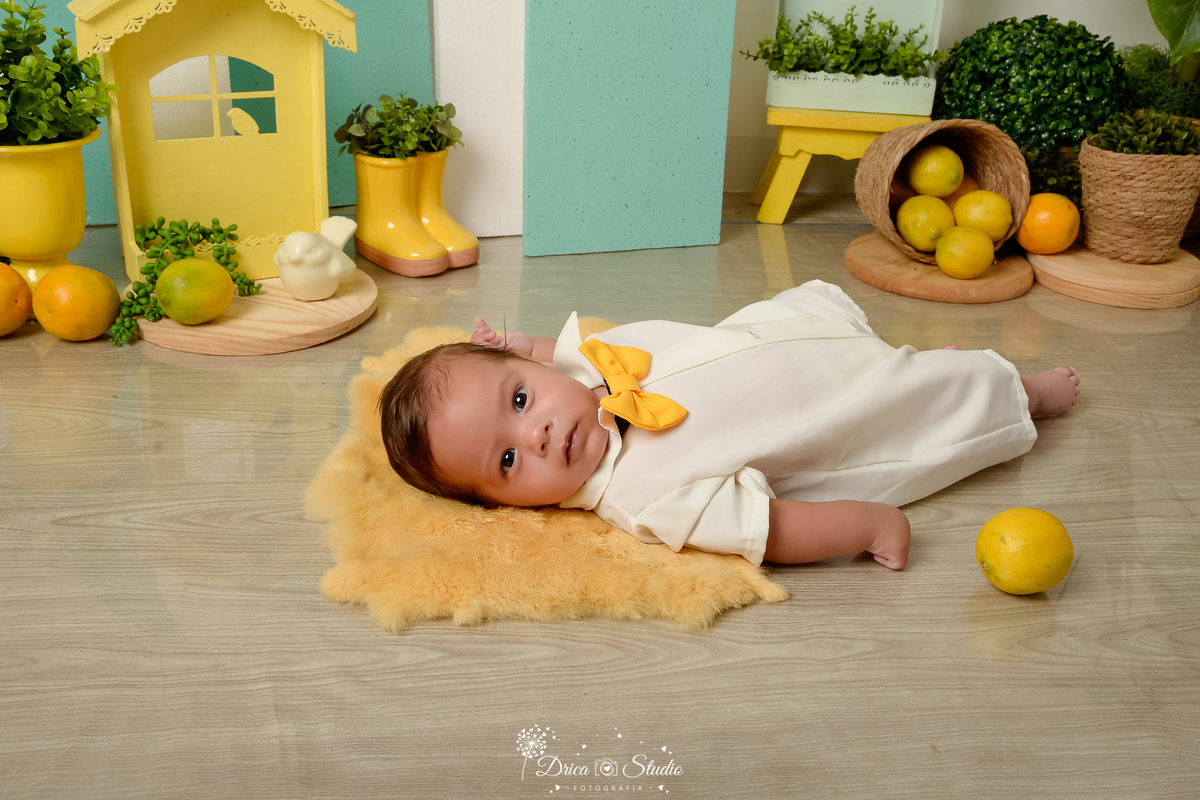 drica studio-ensaio fotográfico infantil - acompanhamento mensal - tema limãozinho - bebê deitado sobre tapete felpudo - cesto de palha com limões - casinha amarela - botinha amarela com planta - passarinho branco - vaso amarelo - piso amadeirado