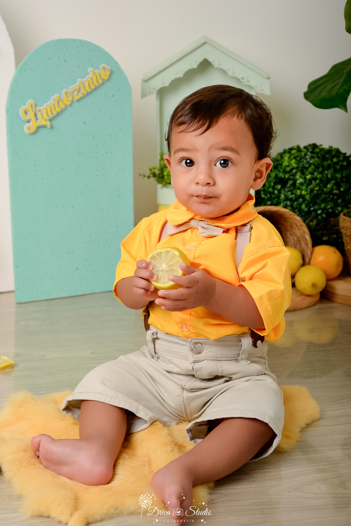 drica studio-ensaio fotográfico infantil - acompanhamento mensal - tema limãozinho - bebê sentado no chão segurando limão, ele veste camisa amarela e short bege - cesto com limões - painel verde - piso amadeirado - 