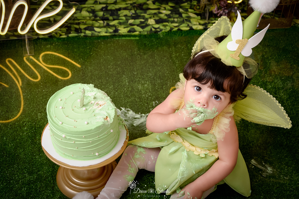 DRICA STUDIO - ENSAIO FOTOGRAFICO INFANTIL - SMASH THE CAKE - TINKER BELL - XINGUARA - BEBÊ VESTIDA DE TINKER BELL, SENTADA AO LADO DE BOLO, COM ROSTO SUJO DE GLACÊ - PISO COM GRAMA SINTETICA - ONE DE LED