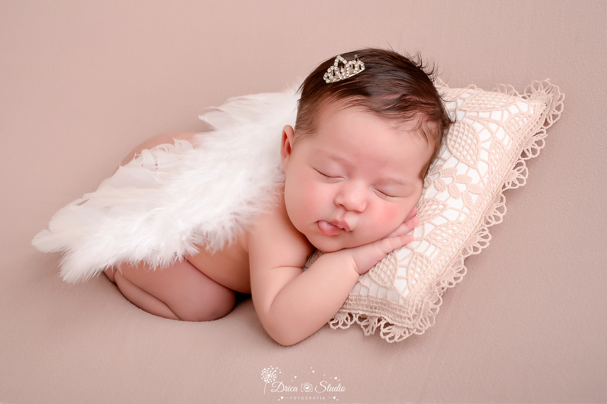 drica studio - ensaio fotografico infantil- recém-nascido- newborn-xinguara-pará-bebê deitada sobre superficie macia com rosto sobre trevesseiro revestido com crochê, sobre sua costa tem uma asa branca e uma coroinha pequena na cabeça - fundo neutro