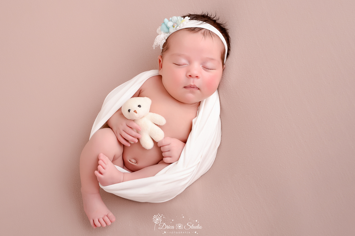 drica studio - ensaio fotografico infantil- recém-nascido- newborn-xinguara-pará- bebê envolvido em manta branca tipo casulo, segurando ursinho de pelucia branco e usando tiara branca com flor azul na cabeça - fundo neutro