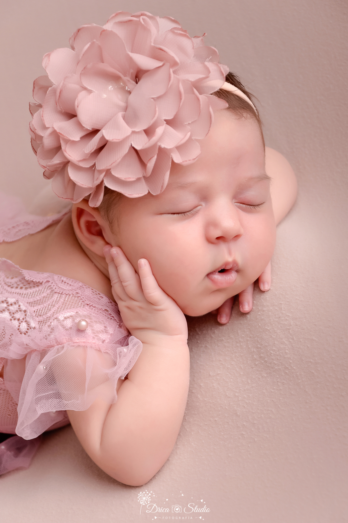 drica studio - ensaio fotografico infantil- recém-nascido- newborn-xinguara-pará-closer de bebê recém-nascida com a mão no rosto, ela usa bory rose e laço de flor na cabeça, está deitada sobre superficie marcia na cor bege
