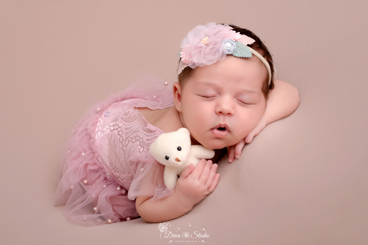 drica studio - ensaio fotografico infantil- recém-nascido- newborn-xinguara-pará-bebê deitada de bruços sobre superficie marcia, veste bory rosa com laço de flor rosa na cabeça, segurando um ursinho branco
