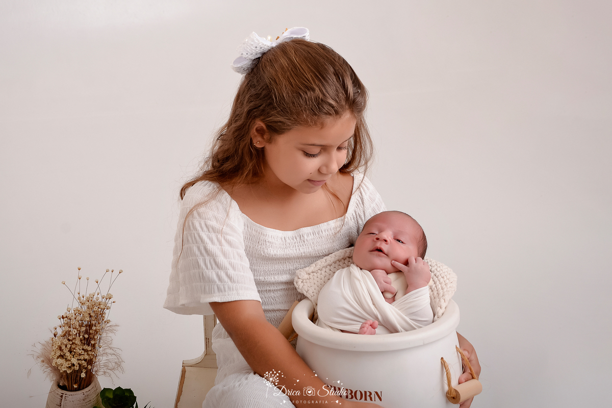drica studio - ensaio fotografico infantil - ensaio recém-nascido - newborn - menina segurando baldinho de madeira branco com irmãozinho recém-nascido dentro - arranjo de flores desidratadas - fundo branco
