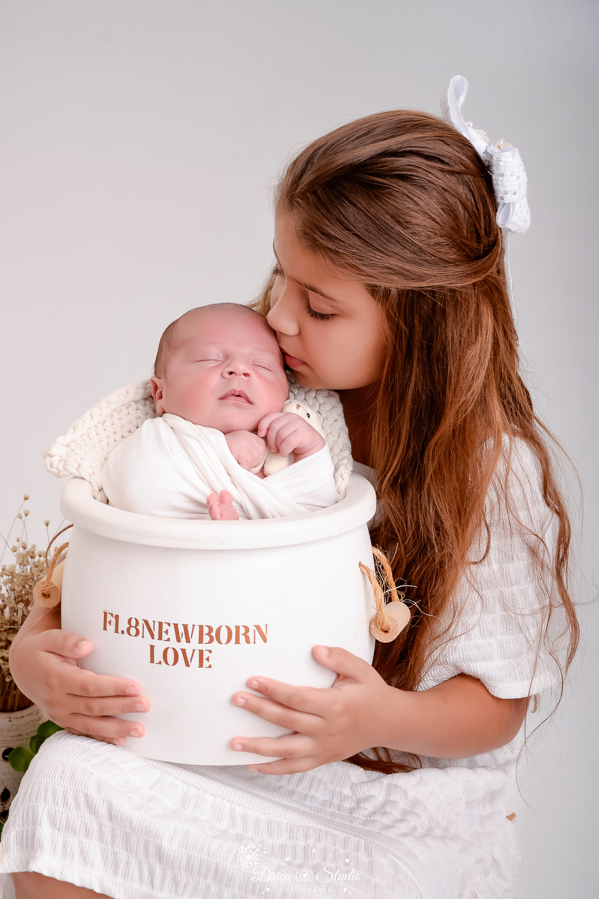 drica studio - ensaio fotografico infantil - ensaio recém-nascido - newborn - menina segurando baldinho com irmãozinho dentro, ela beija a cabecinha do bebê, usa vestido branco e bebê está enrolado em manta branca