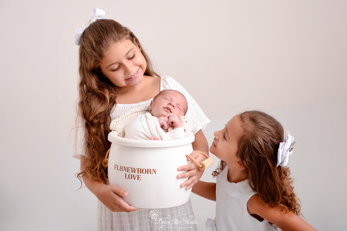 drica studio - ensaio fotografico infantil - ensaio recém-nascido - newborn - irmãs mais velhas segurando baldinho de madeira com irmãozinho recém-nascido dentro - elas usam vestidos branco, o bebê está enrolado em manta branca - fundo neutro