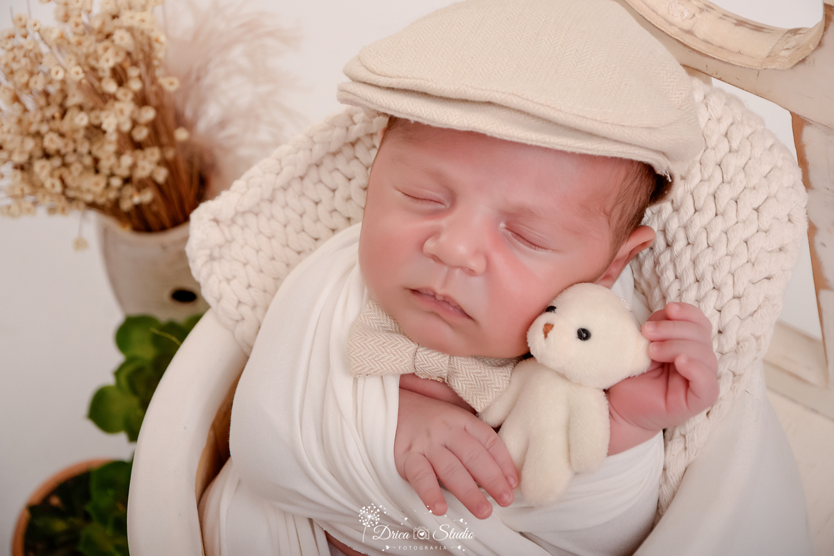 drica studio - ensaio fotografico infantil - ensaio recém-nascido - newborn - bebê enrolado em manta tipo casulo dentro de baldinho de madeira branco - ursinho de pelúcia branco e boina bege clara na cabeça - cadeira de madeira - vaso com flores seca