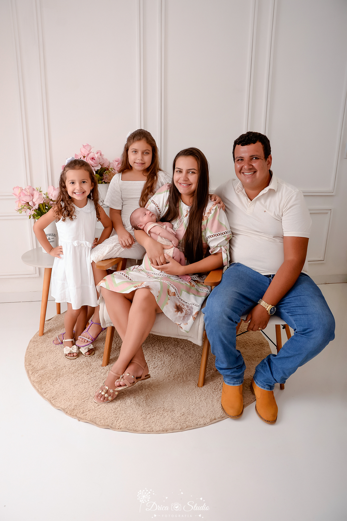drica studio - ensaio fotografico infantil - ensaio recém-nascido - newbon-família em pose para foto mostrando o bebê recém-nascido-as meninas usam vestido branco, a mãe vestido florido e o pai camiseta branca com calça jeans-fundo branco com boiserie

