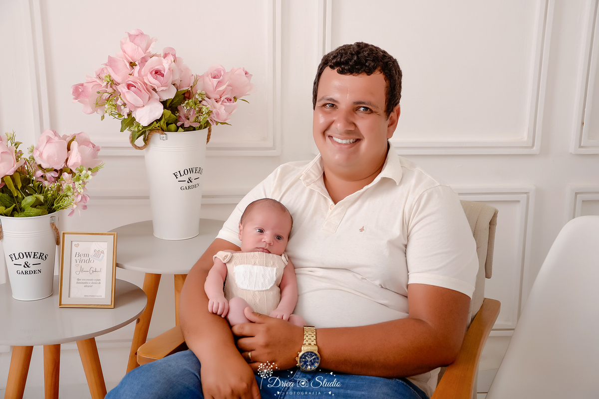 drica studio - ensaio fotografico infantil - ensaio recém-nascido - newborn - pai sentado em poltrona com bebê recém-nascido no colo - fundo branco com boiserie - mesinha cinza com detalhes em madeira - vaso branco com flores rosa - 