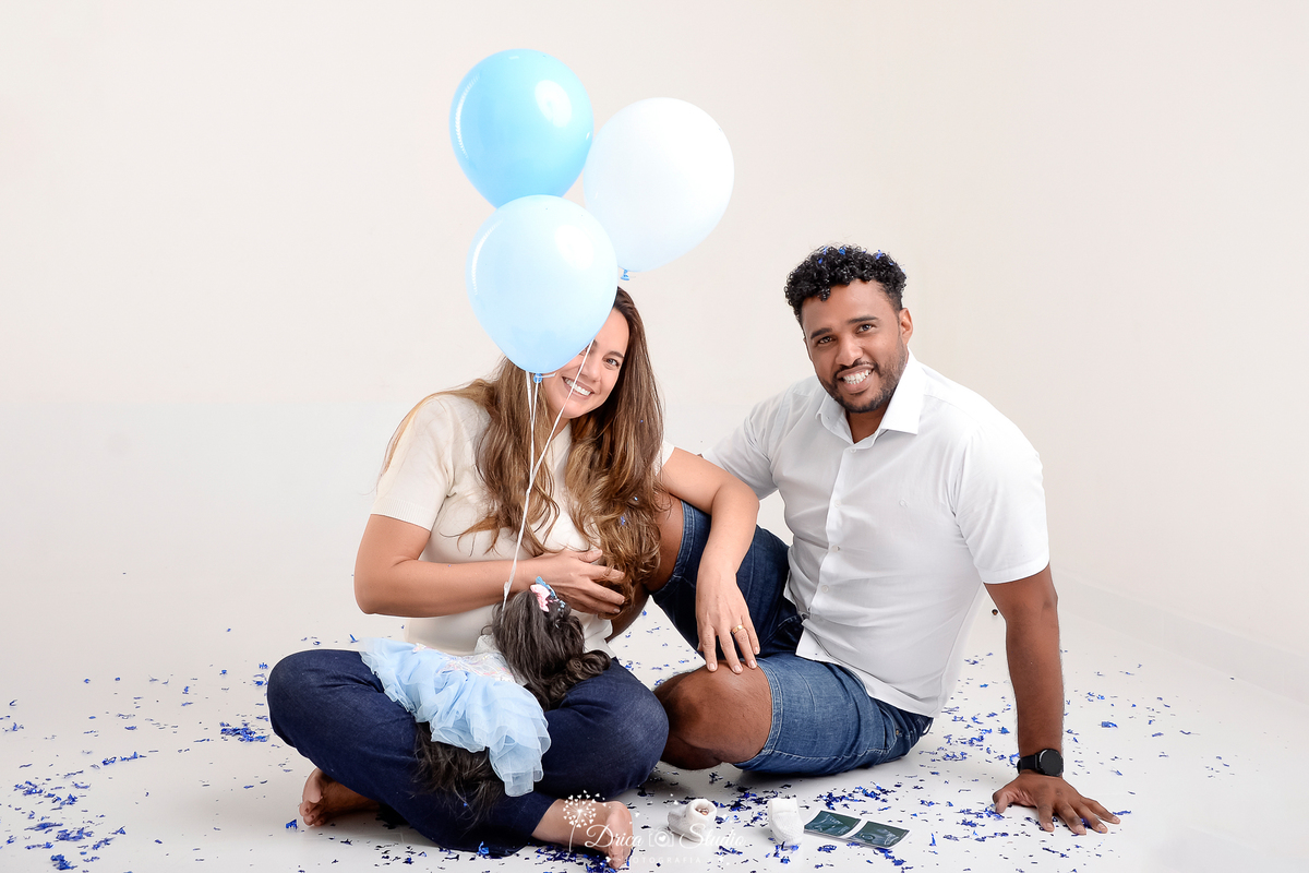 drica studio -ensaio fotografico adulto - chá revelação - sexo do bebê - xinguara - pará - casal sentado no chão, em sua volta confetes azuis, balão azuis preso a cachorrinha preta - fundo clean