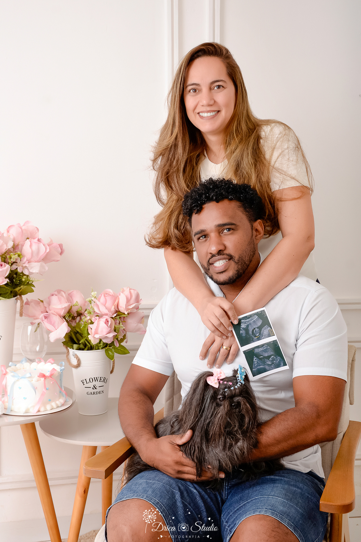 drica studio -ensaio fotografico adulto - chá revelação - sexo do bebê - xinguara - pará - homem sentado em poltrona com cachorrinha preta no colo e esposa em pé atrás do esposo com braços envolvido a seu pescoço segurando um ultrassonografia-fundo branco