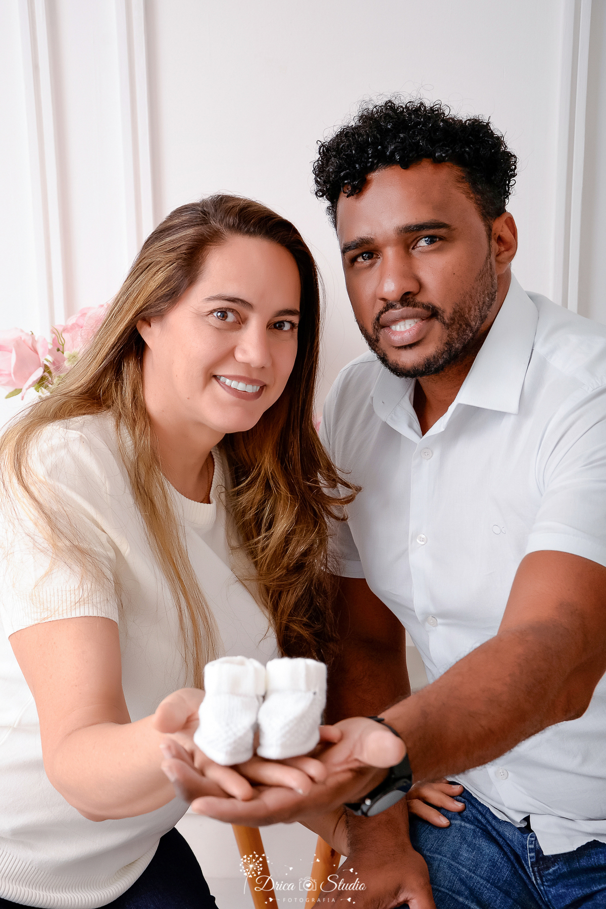 drica studio -ensaio fotografico adulto - chá revelação - sexo do bebê - xinguara - pará - casal com as mãos estendida mostrando sapatinho de bebê branco - mulher veste blusa bege clara e esposo camisa branca - fundo claro