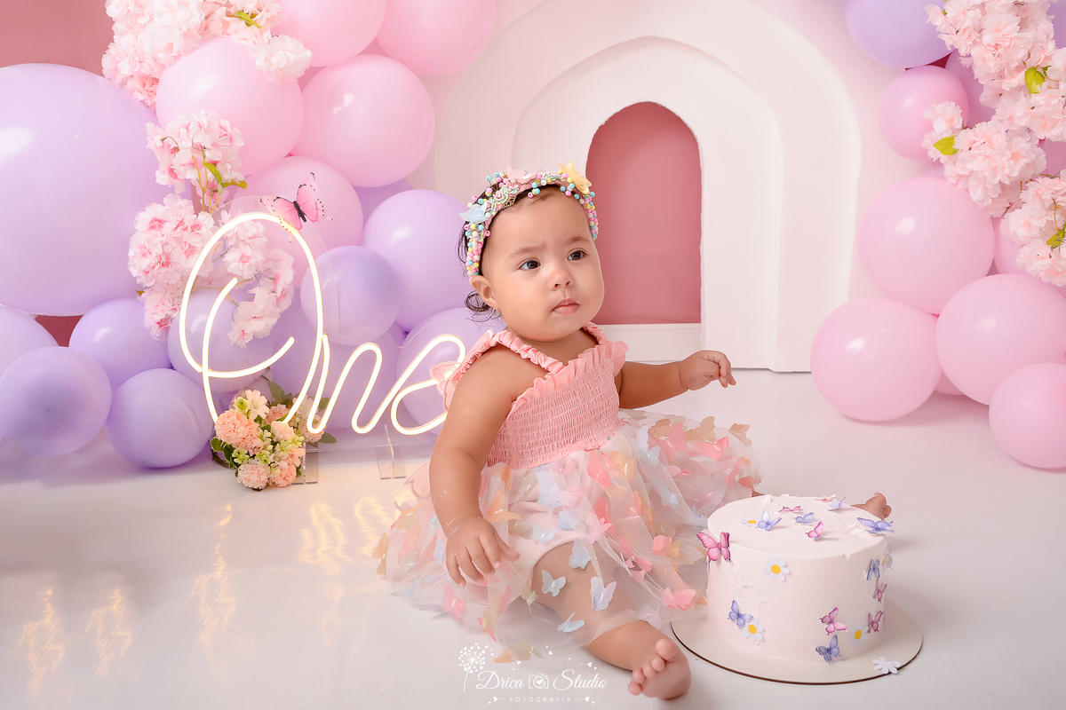 drica studio - ensaio fotográfico infantil - smash the cake - tema jardim de borboletas - xinguara - pará- bebê sentada no chão com bolo a sua frente- one de led - balões rosa e lilas - flores rosa - painel branco - fundo rose - vestido  e tiara rosa- 