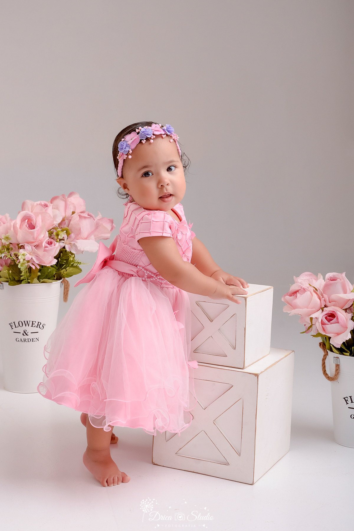 drica studio - ensaio fotográfico infantil - smash the cake - tema jardim de borboletas - xinguara - pará- bebê usando vestido rosa com tule em pé se apoiando em caixotes de madeira branco - vaso branco com flores rosa - fundo neutro